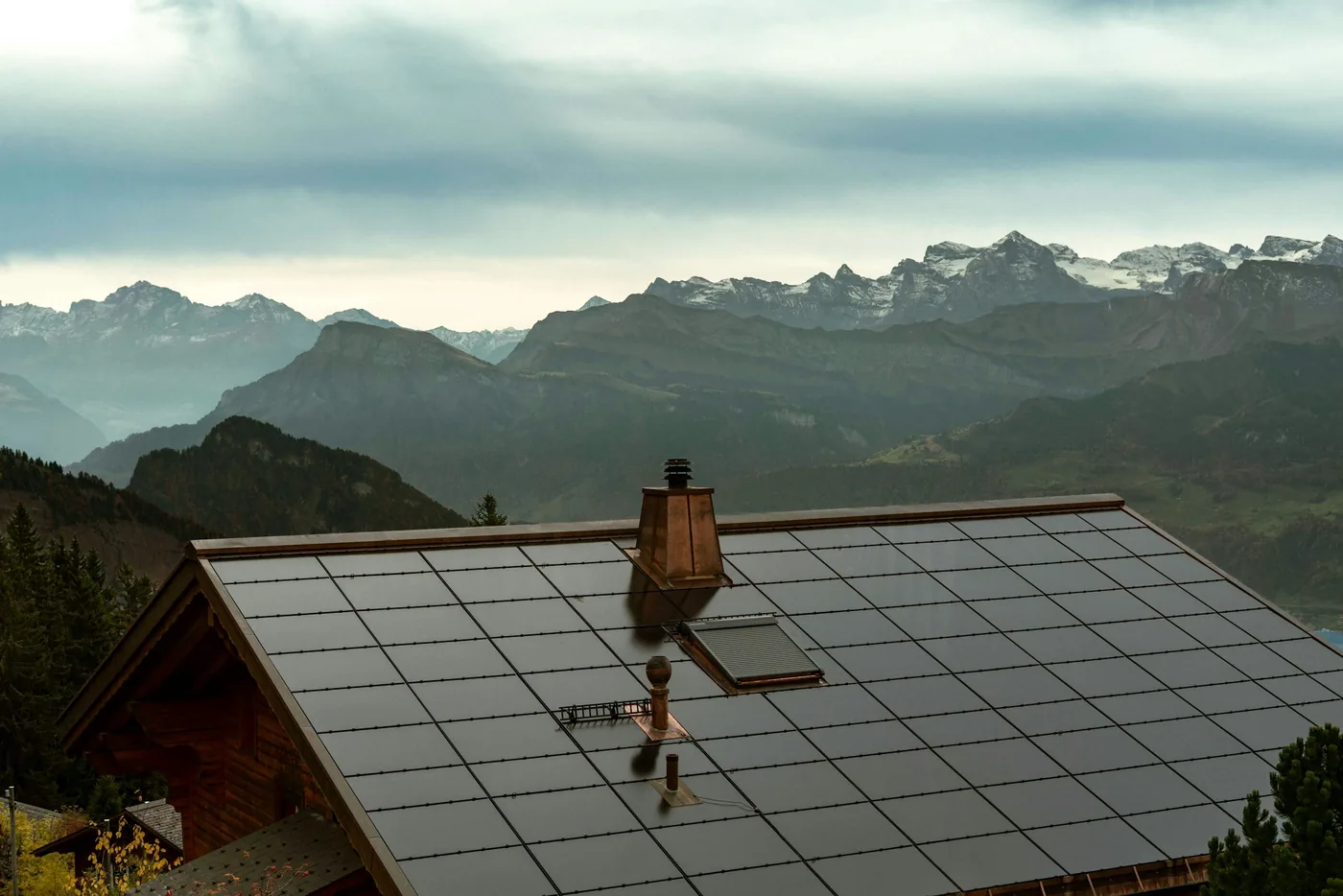 Dach eines Holzhauses vollflächig mit dunklen Solarmodulen gedeckt, im Hintergrund Alpengipfel (Runding, Landkreis Cham)