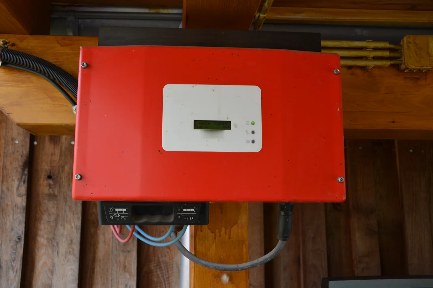 Roter Wechselrichter mit Display an einem Holzbalken, darunter die Kabelanschlüsse (Espenau, Landkreis Kassel)