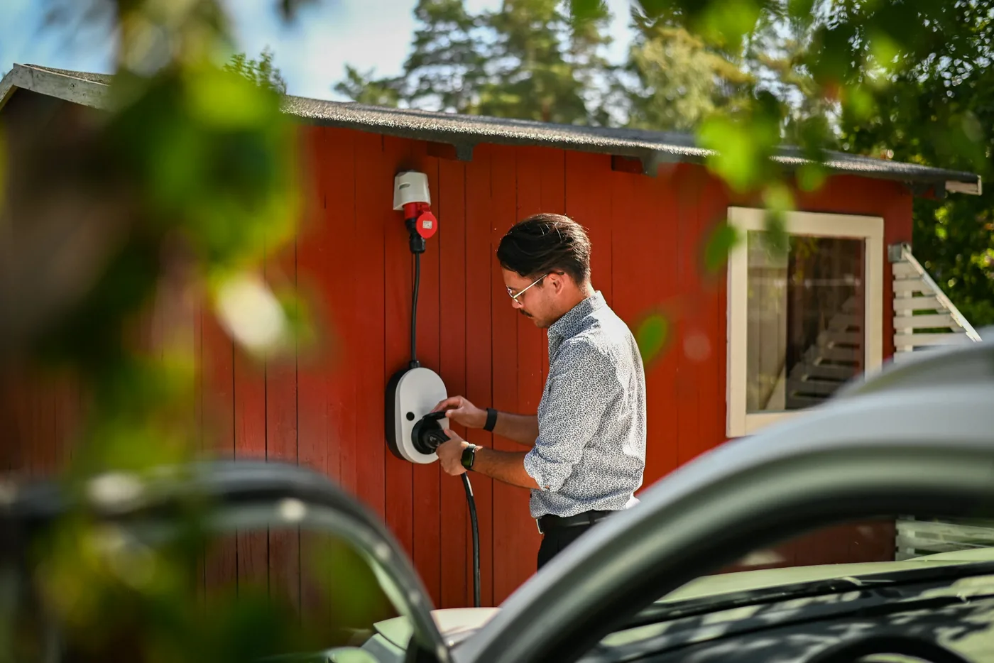 Mann am Ladestecker einer Wandladestation an einem roten Holzhaus, vorne das Elektroauto